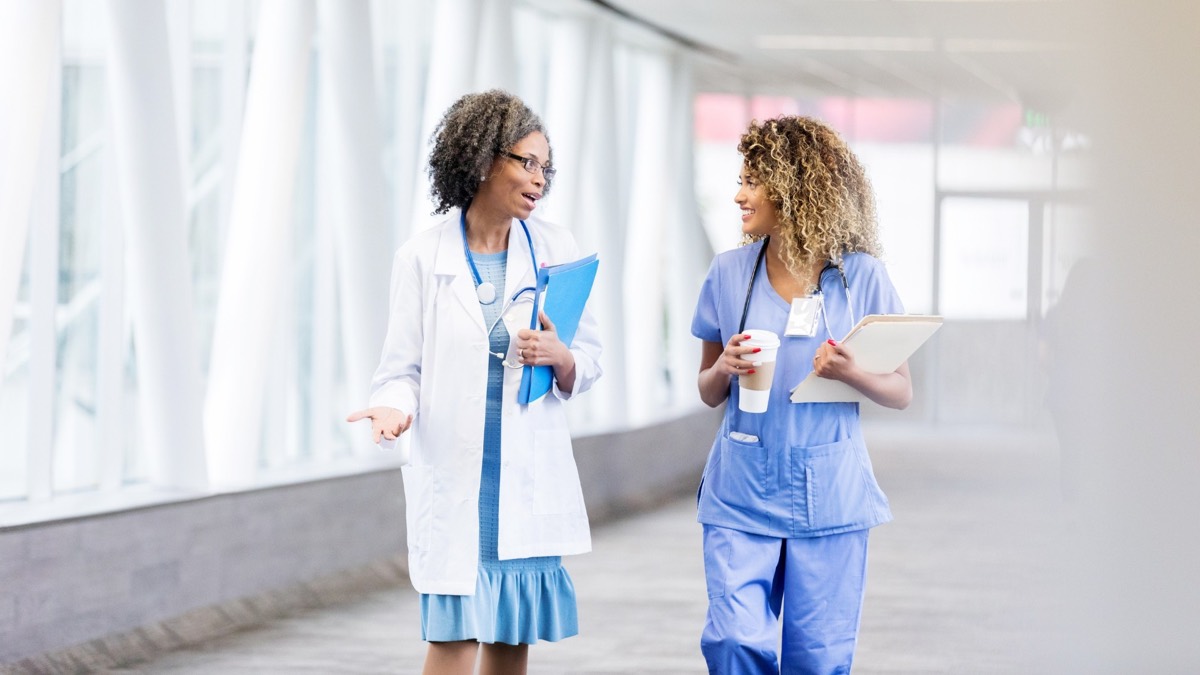 Eine Ärztin und eine Pflegekraft gehen mit einem Kaffee in der Hand über den Klinikflur